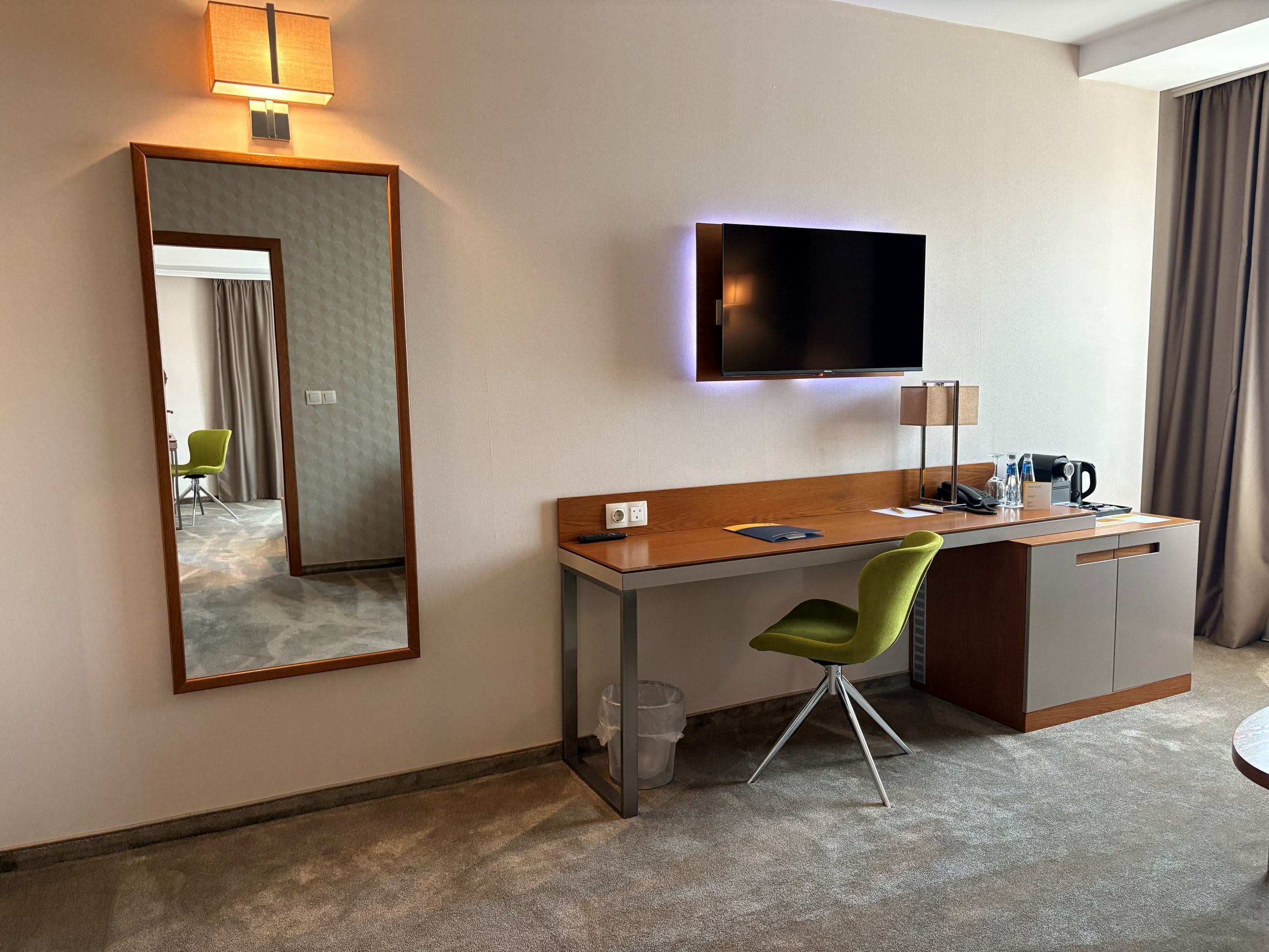 Habitación de hotel con escritorio, una silla verde, televisión de pantalla plana, espejo, lámpara y cafetera.