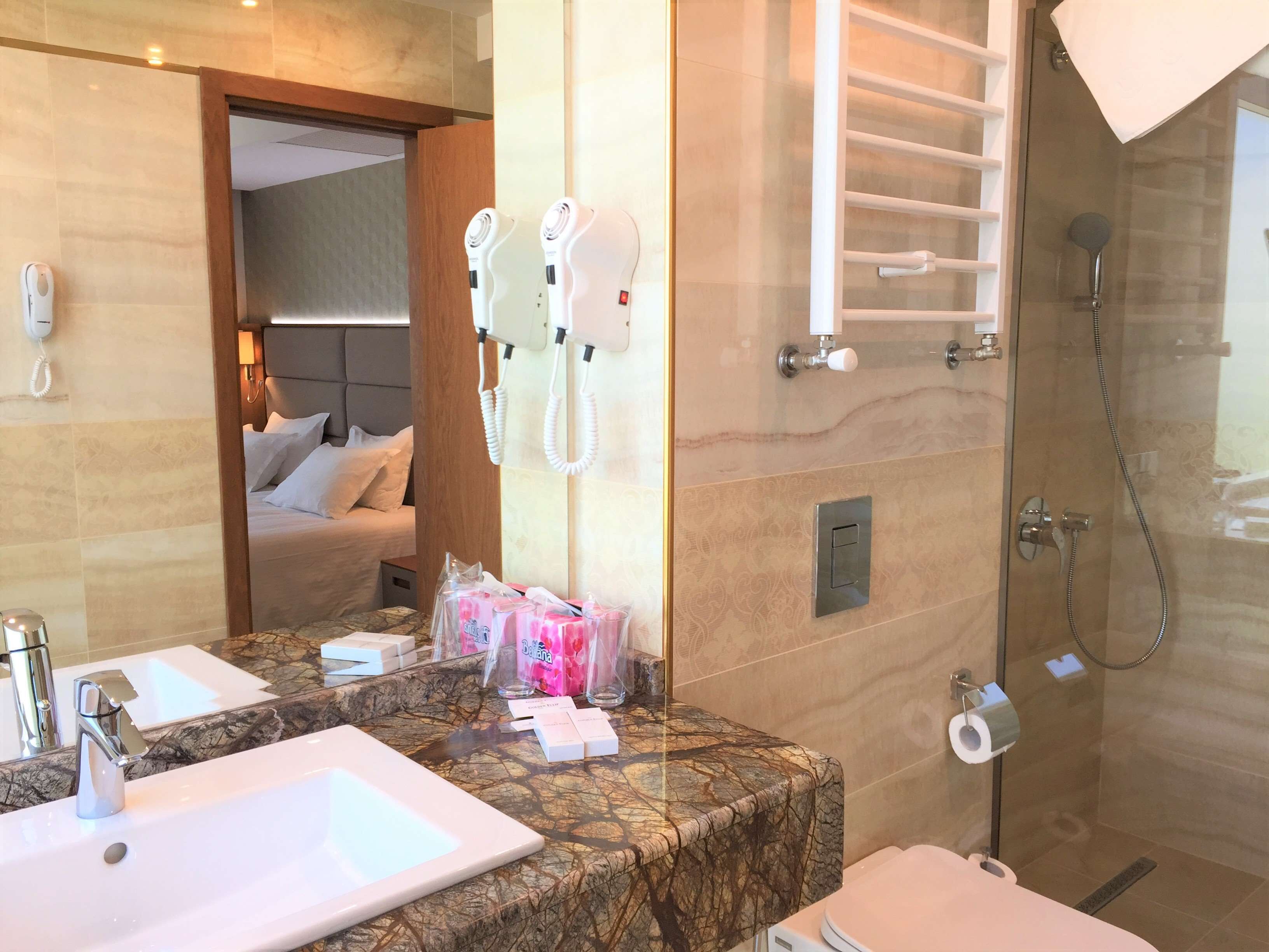 A hotel bathroom with a marble countertop, two sinks, a shower, and a bed visible through an open door.