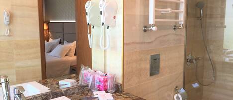 A hotel bathroom with a marble countertop, two sinks, a shower, and a bed visible through an open door.
