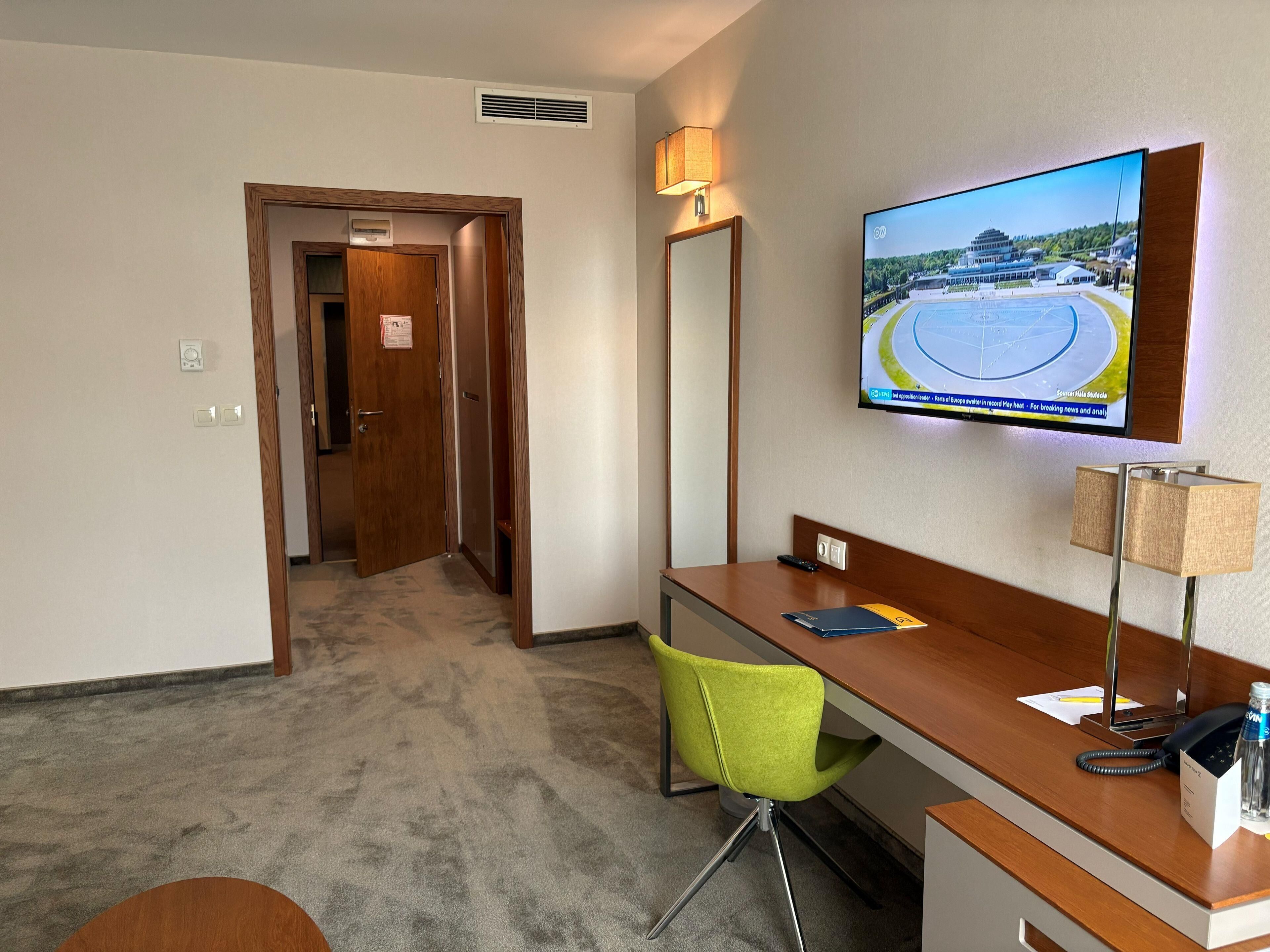 Een hotelkamer met een bureau, een groene stoel, een aan de muur bevestigde televisie en een deur naar een andere kamer.