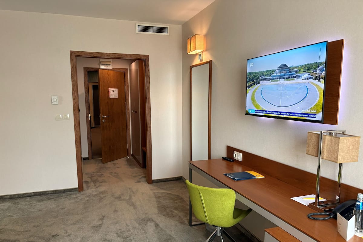 A hotel room with a desk, a green chair, a television mounted on the wall, and a door leading to another room.