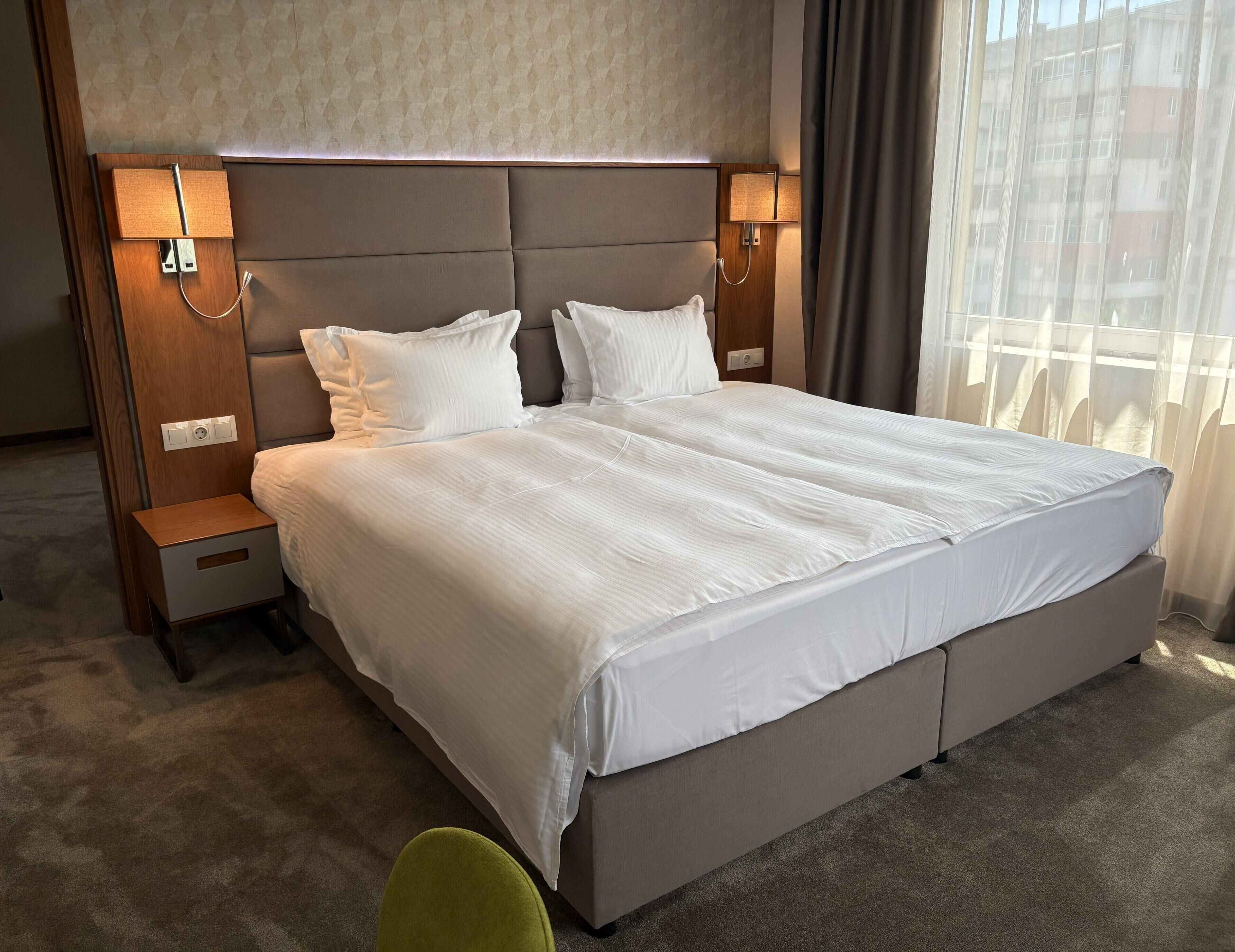 Habitación de hotel con una cama grande, mesitas de noche, una lámpara y vistas a edificios a través de la ventana.