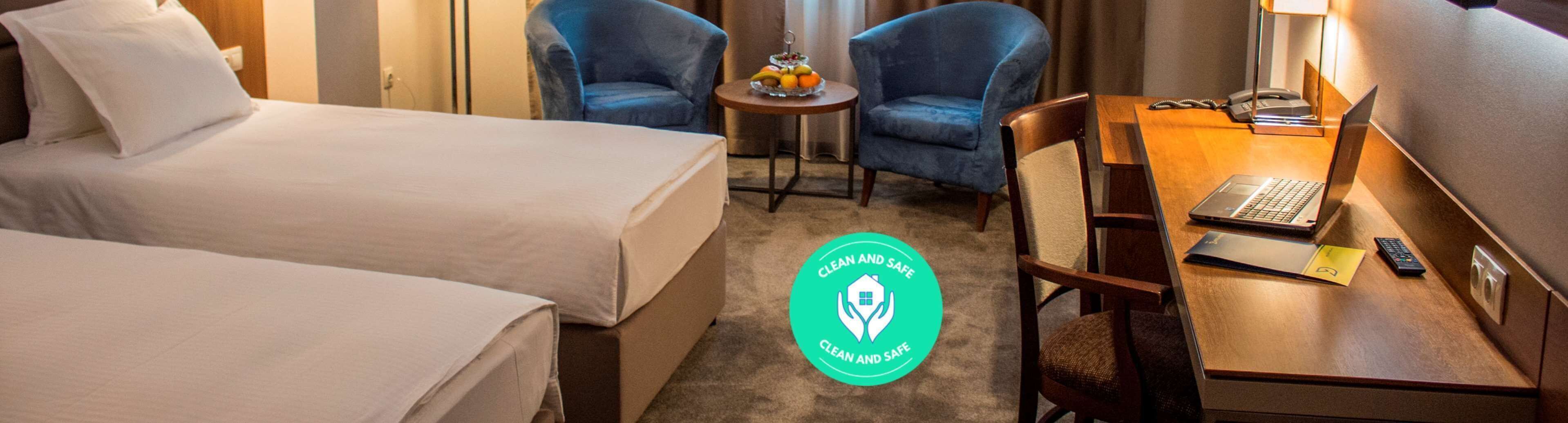 Ein Hotelzimmer mit zwei Betten, einem Schreibtisch mit einem Laptop, einem Stuhl, einem kleinen Tisch mit Obst und dem Logo 'Clean and Safe'.