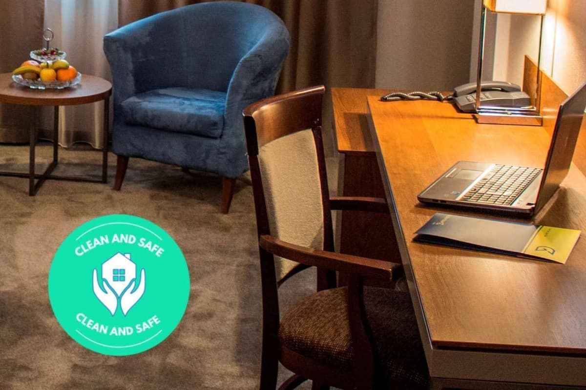 A hotel room with two beds, a desk with a laptop, a chair, a small table with fruit, and a 'Clean and Safe' logo.