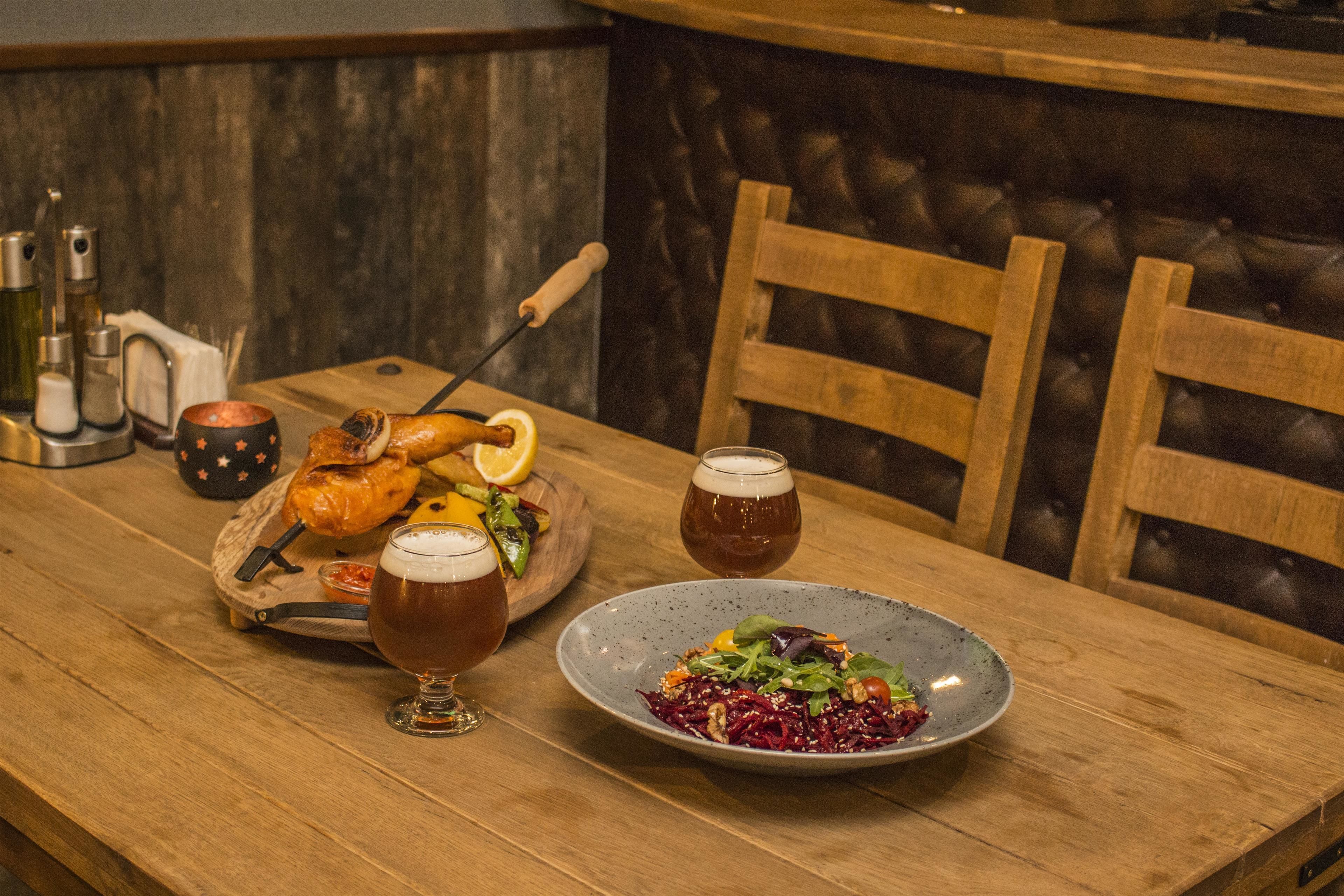 A wooden dining table with a roasted chicken, a plate of salad, and two glasses of beer.