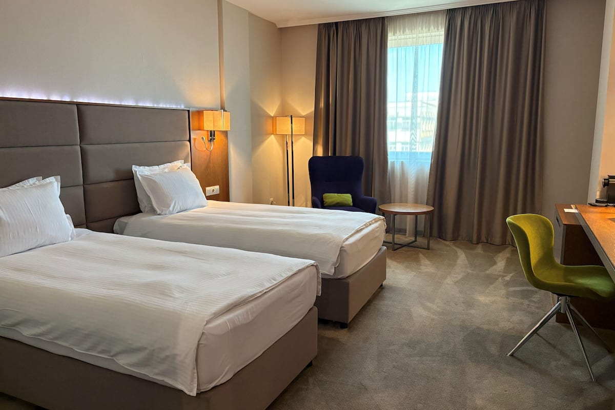 A hotel room with two beds, a desk, a chair, a lamp, and a view of the outside.