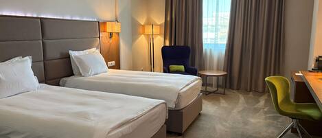 Habitación de hotel con dos camas, un escritorio, una silla, una lámpara y vistas al exterior.