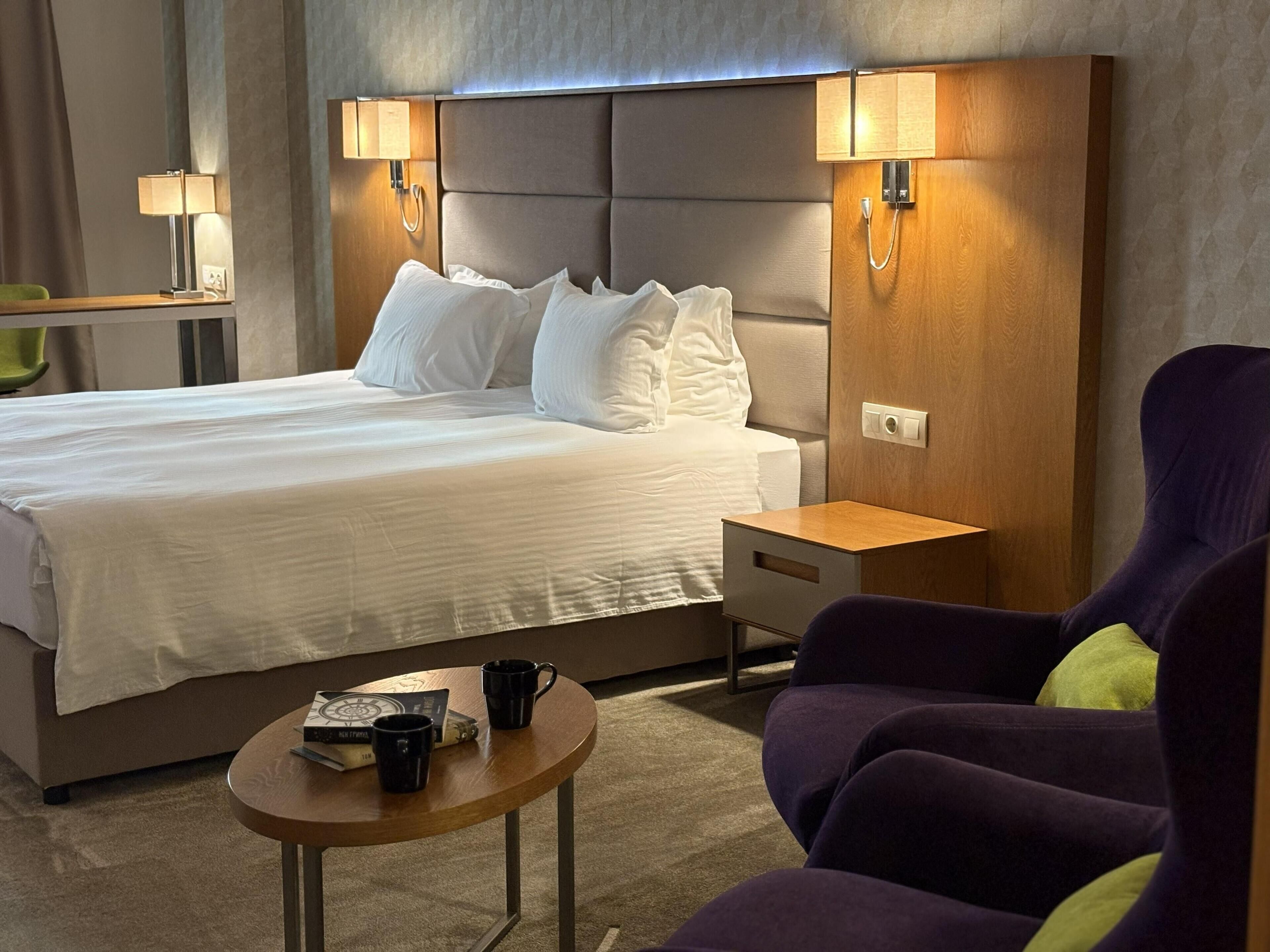 Une chambre d’hôtel avec un lit, des lampes de chevet, une petite table avec un livre et une tasse, et un fauteuil violet.