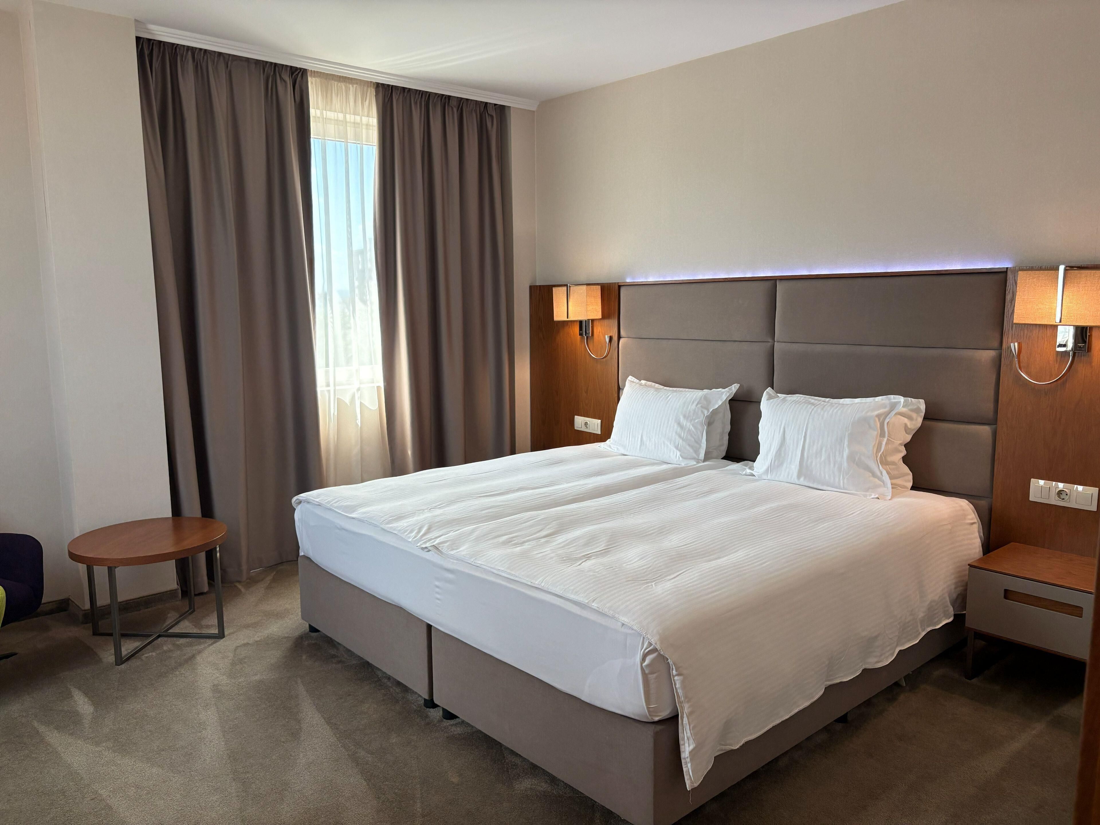 A hotel room with a large bed, two bedside tables, a small round table, and a window with curtains.