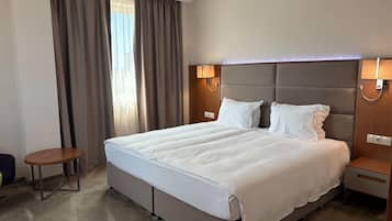 A hotel room with a large bed, two bedside tables, a small round table, and a window with curtains.