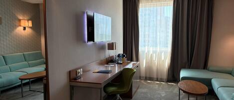 Ein Hotelzimmer mit einer Couch, einem Schreibtisch, einem Stuhl, einem Spiegel, einem Fernseher und einem Fenster mit Vorhängen.