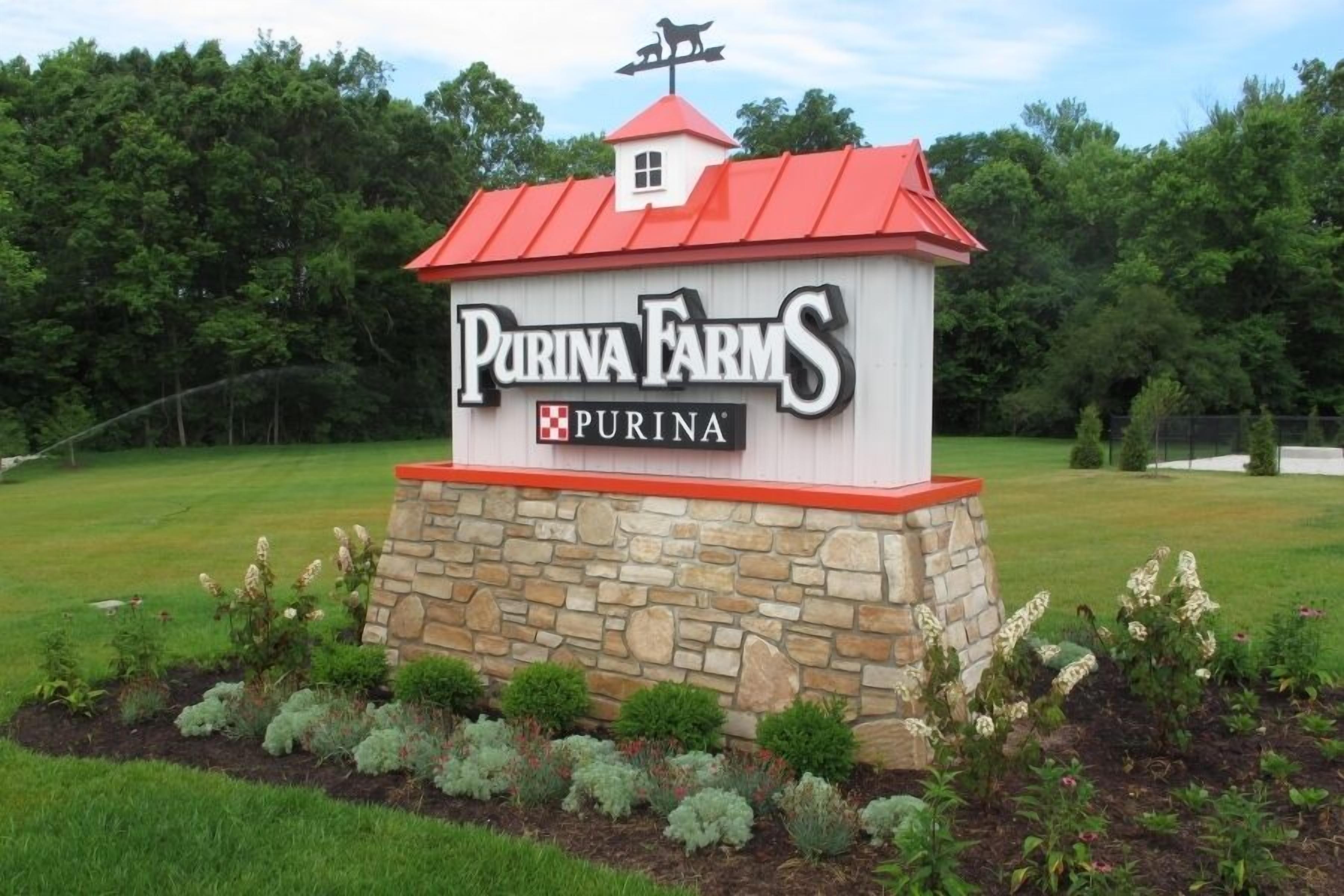 Señal de Purina Farms con base de piedra y techo rojo, rodeada de jardines paisajísticos.