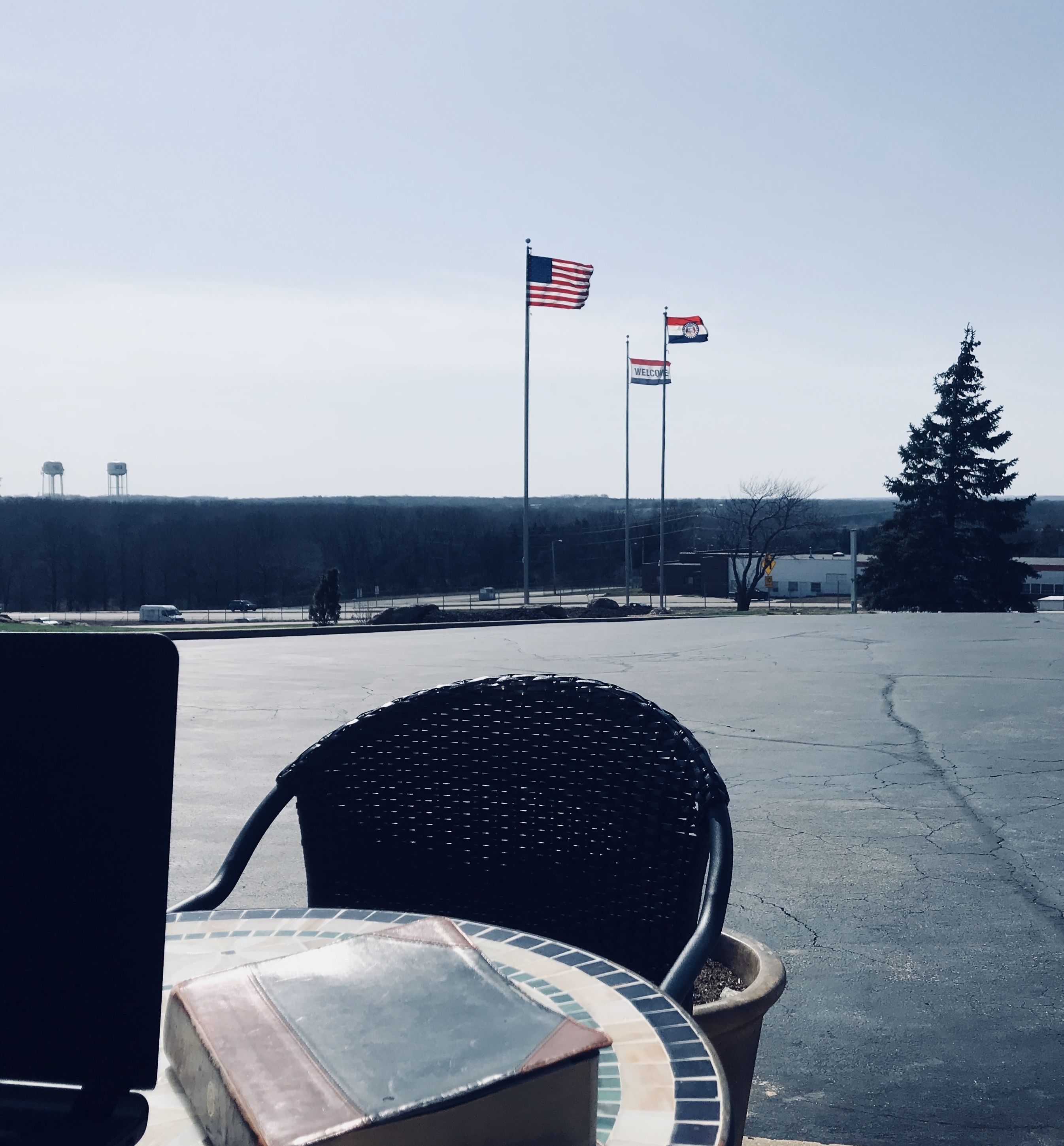 Ein Parkplatz mit einer Reihe von Flaggen, darunter die US-amerikanische Flagge und eine Flagge mit einem 'Welcome'-Schild. Im Vordergrund stehen ein Stuhl und ein Tisch.