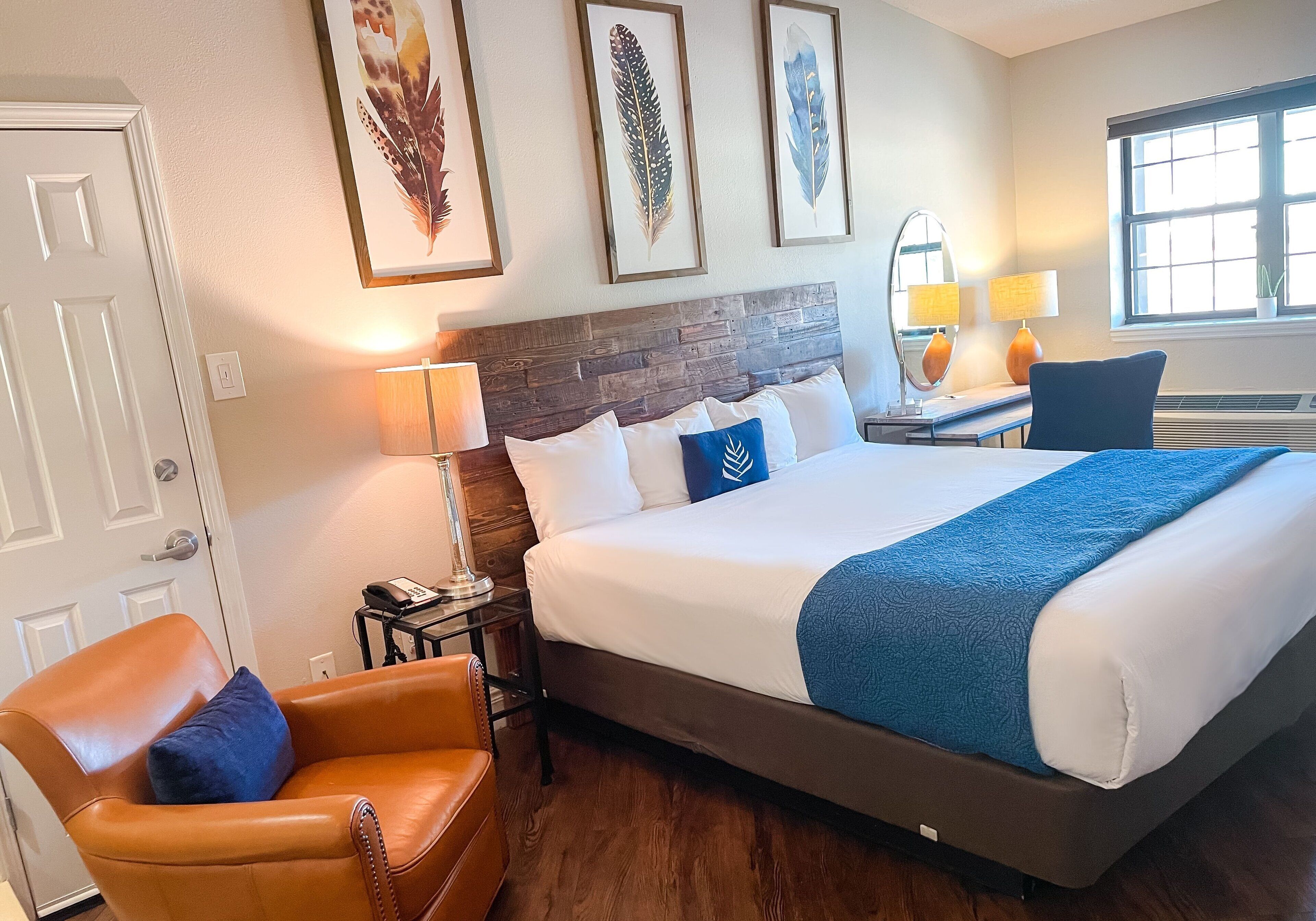 A hotel room with a large bed, a brown leather chair, a desk with a lamp, and feather wall art.