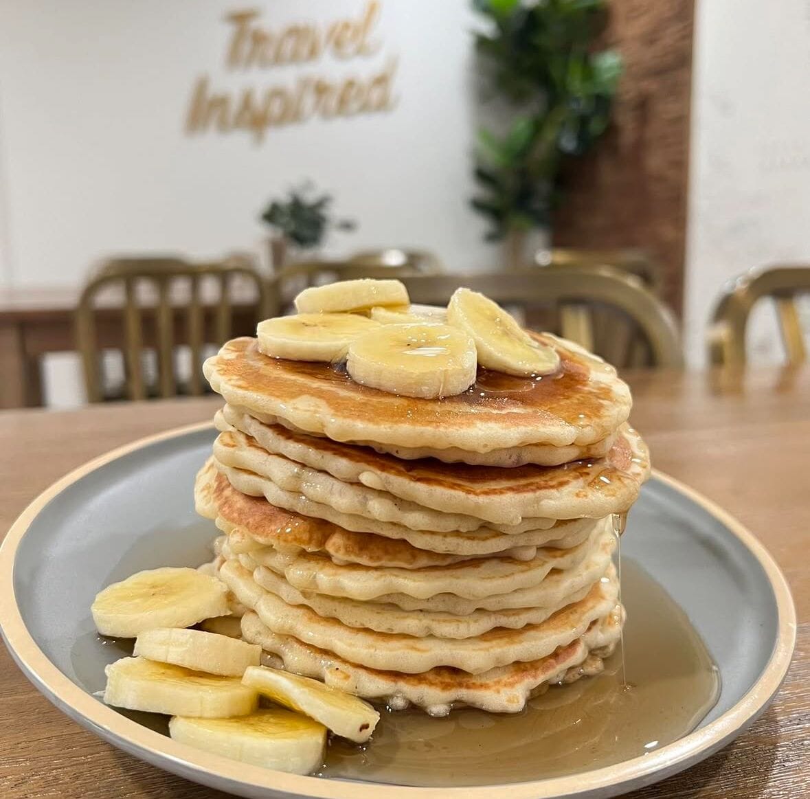 Una pila de tortitas con rodajas de plátano sobre un plato, con un cartel que dice 'Travel Inspired' al fondo.