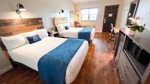 Habitación de hotel con dos camas, piso de madera, un escritorio con silla, microondas y una planta en maceta.