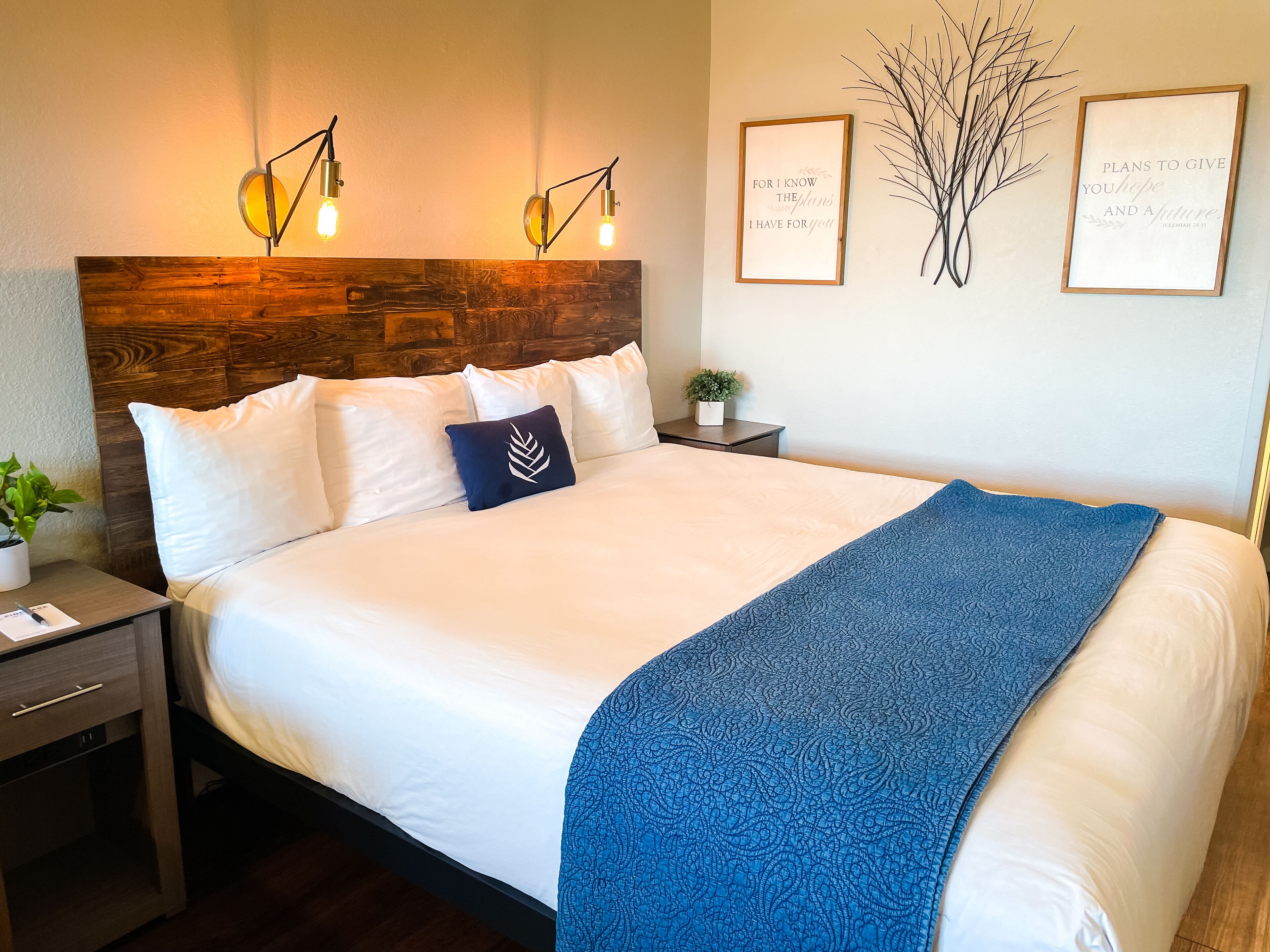 Een net opgemaakt bed met witte lakens en een blauwe plaid, twee nachtkastjes met lampen, een kleine potplant en twee ingelijste inspirerende citaten aan de muur.