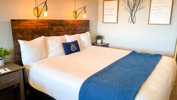 Een net opgemaakt bed met witte lakens en een blauwe plaid, twee nachtkastjes met lampen, een kleine potplant en twee ingelijste inspirerende citaten aan de muur.