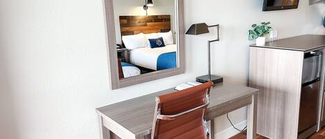 A compact hotel room with a wooden desk, a brown leather chair, a large mirror, a flat-screen TV, a small plant, and a kitchenette with a stainless steel refrigerator.