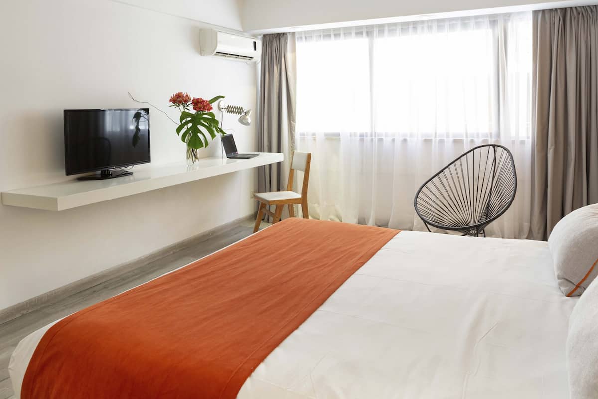 A hotel room with a bed, a TV on a white shelf, a chair, and a window with curtains.