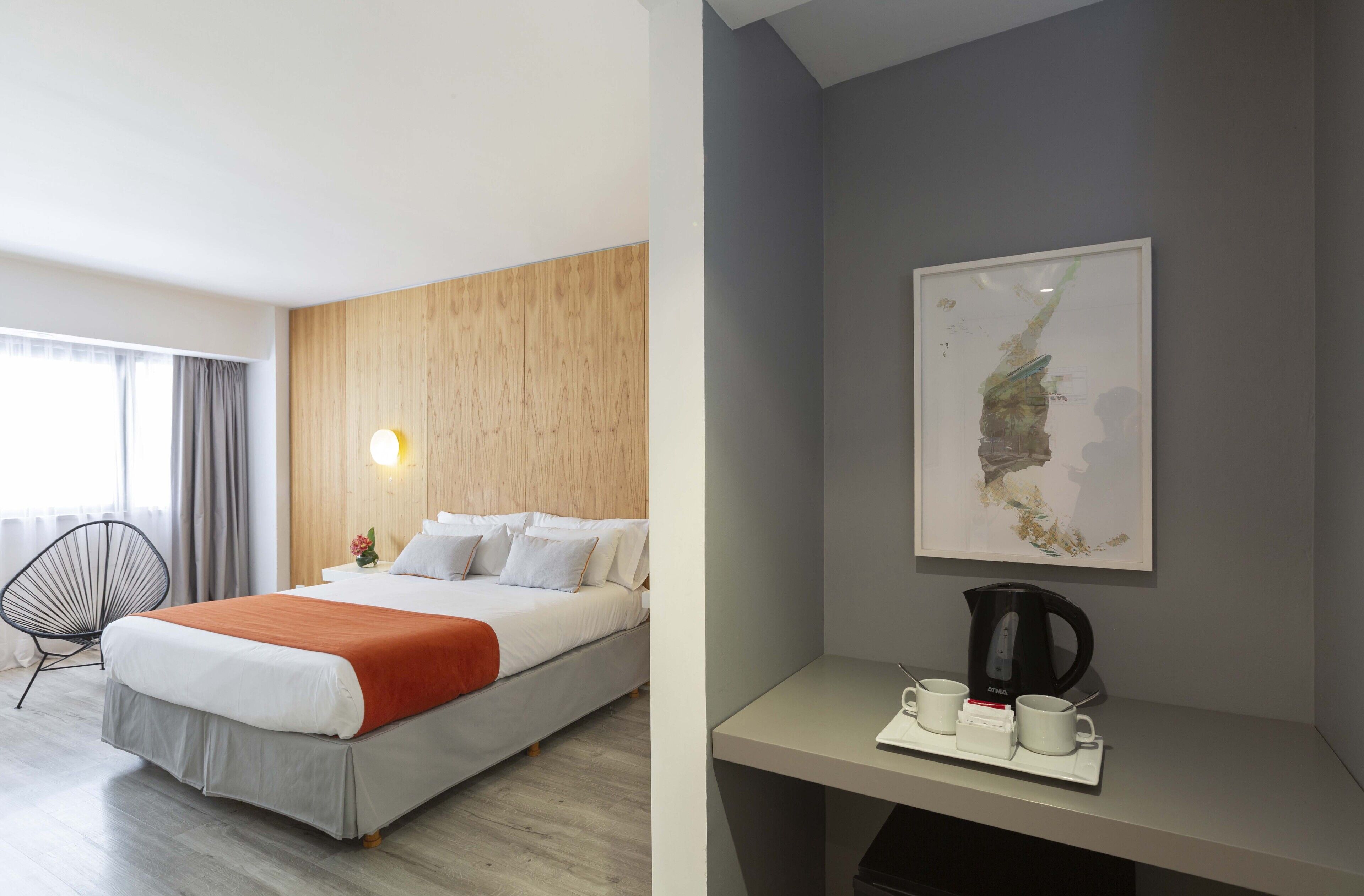 A modern hotel room with a bed, a chair, a coffee station, and a painting on the wall.