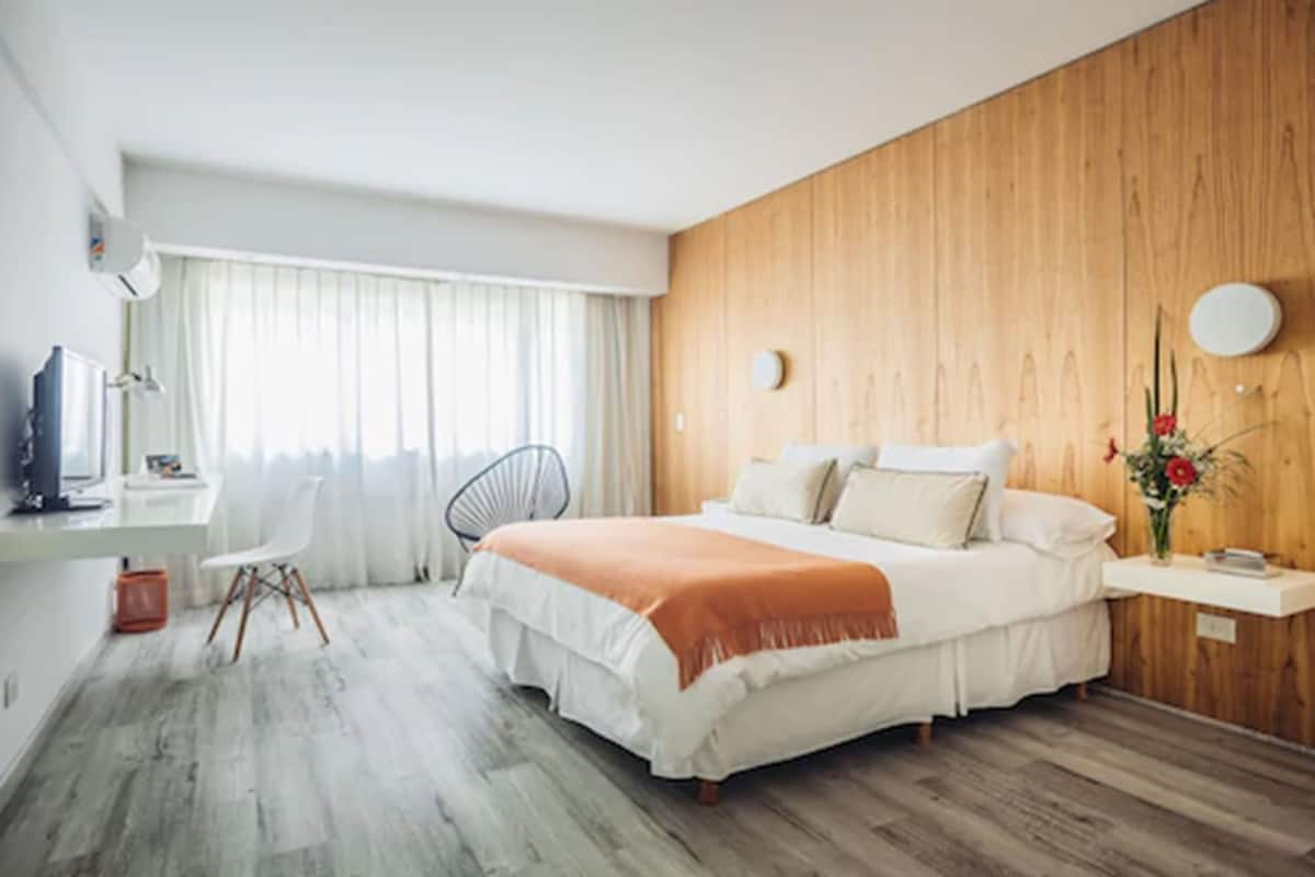 A modern hotel room with a wooden accent wall, a large bed, a desk with a chair, and a window with curtains.