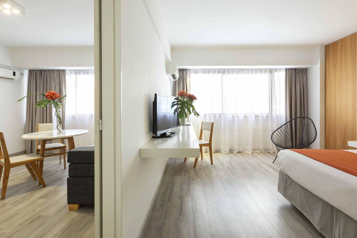 A hotel room with a bed, a desk with a TV, a chair, a dining table with chairs, and a window with curtains.