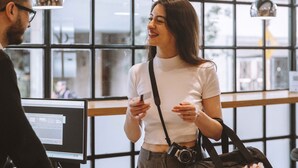 Une personne vêtue d’un haut blanc et d’un pantalon gris, debout dans une pièce dotée de grandes fenêtres et d’un écran d’ordinateur.