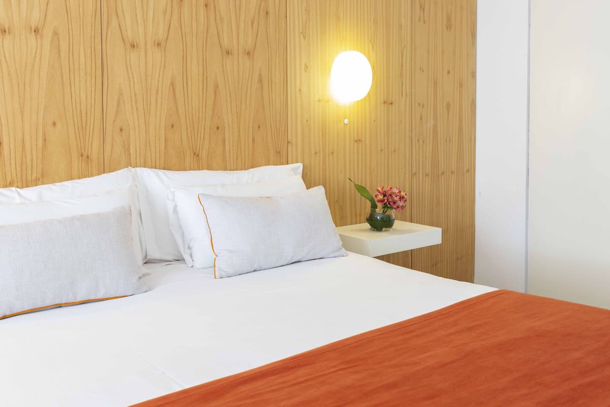 A neatly made bed with white linens and an orange throw blanket, a bedside table with a vase of flowers, and a wall-mounted light fixture.