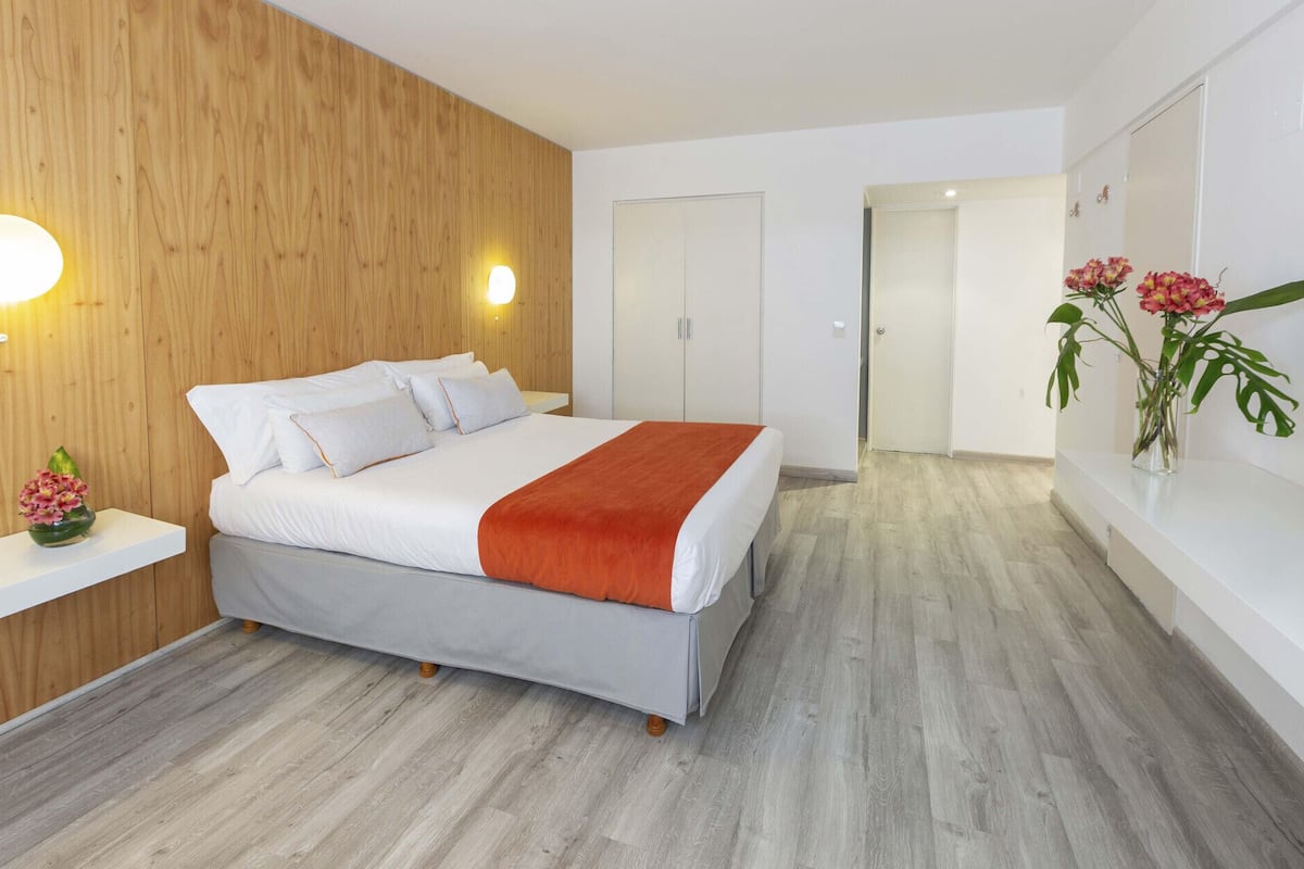 A modern bedroom with a bed, wooden panel wall, white bedside table, and a vase with flowers.