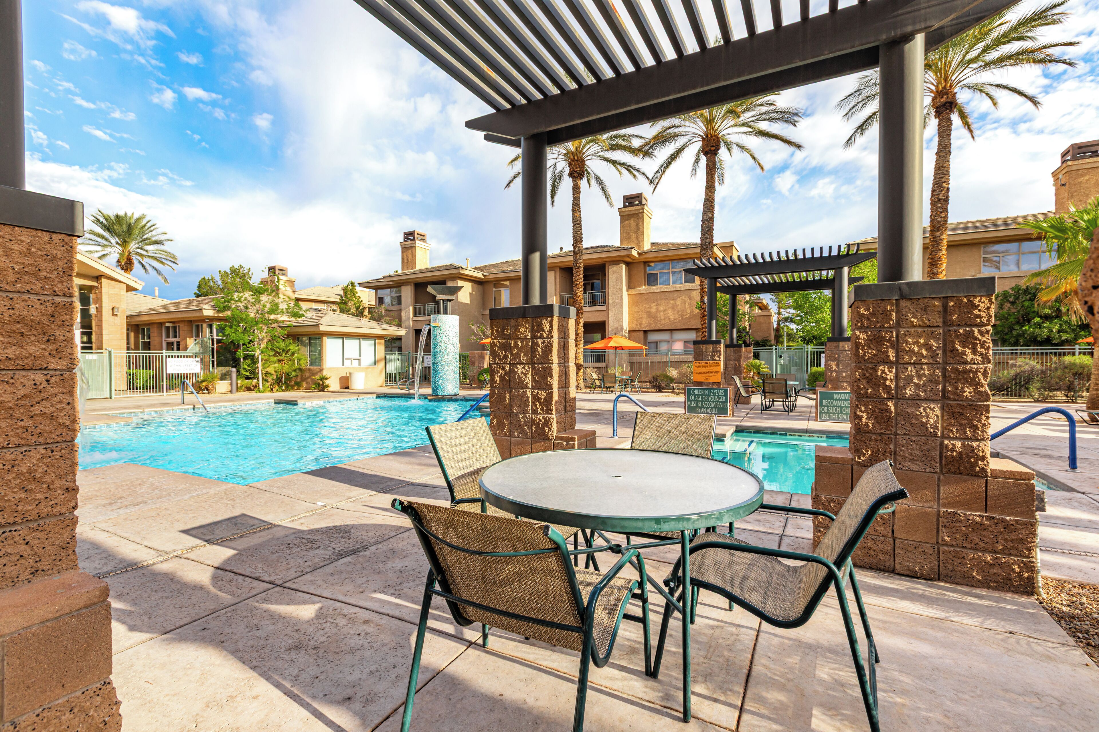A pool area with a table and chairs, palm trees, and a covered seating area.