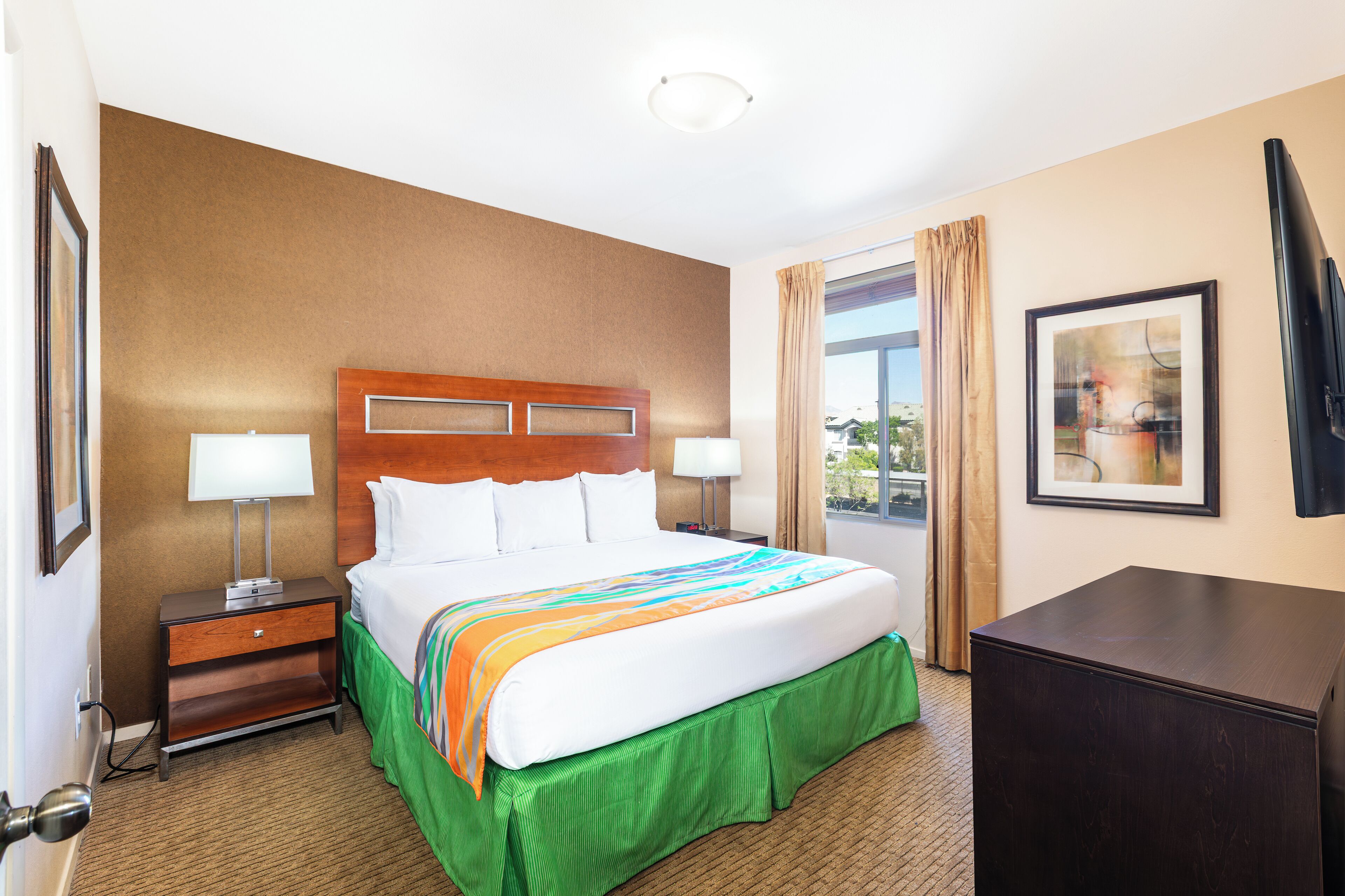 Quarto de hotel com uma cama grande, duas mesas de cabeceira, uma escrivaninha, uma janela com cortinas e um quadro emoldurado na parede.