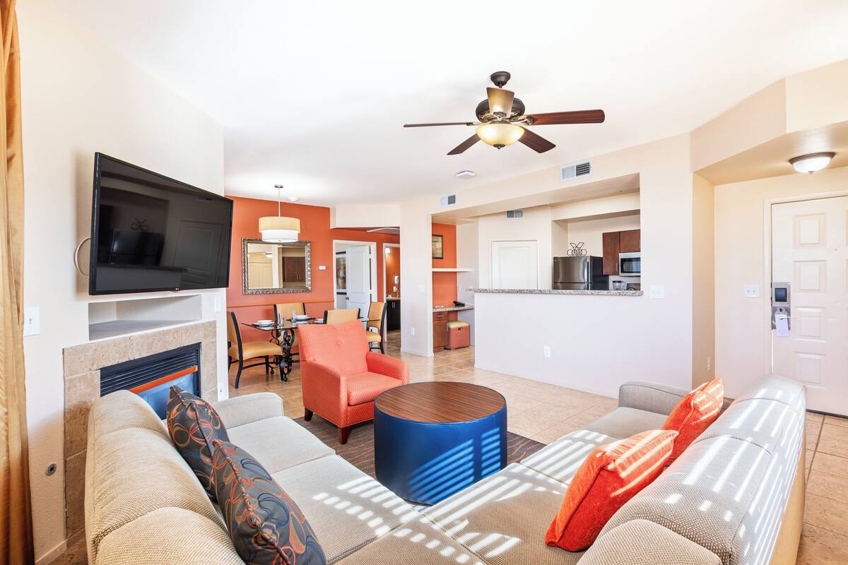 A modern living room with a flat-screen TV, a fireplace, a ceiling fan, and a kitchen area in the background.