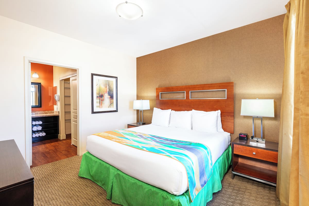 A hotel room with a large bed, a wooden headboard, two bedside tables with lamps, a green skirted bench, and a framed picture on the wall.