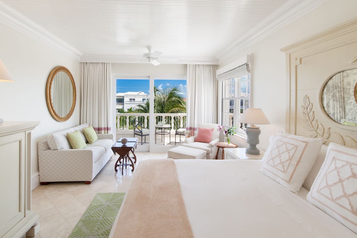 Een ruime slaapkamer met een groot bed, een zithoek met een bank en fauteuils, een balkon met uitzicht op palmbomen, en een plafondventilator.