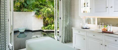 A spa room with a glass-enclosed hot tub, a massage table, and a vanity with toiletries.