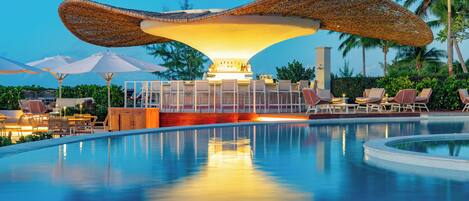 A pool area with lounge chairs and umbrellas, a bar area with a distinctive overhead structure, and a view of palm trees.