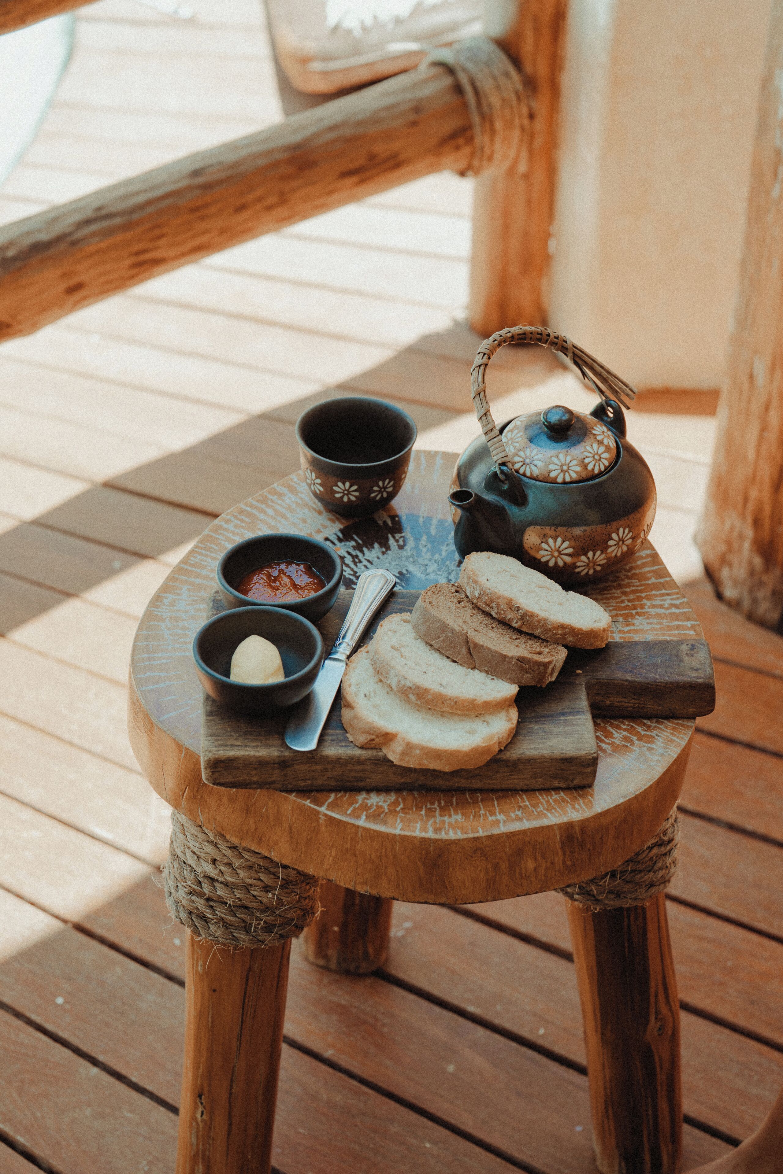 A wooden table with a teapot, two bowls, a knife, and sliced bread on a wooden deck.