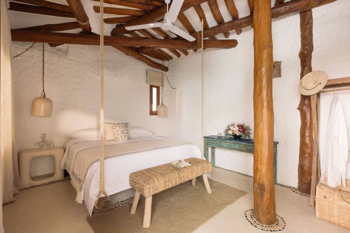 A rustic bedroom with a bed, bench, hanging light, wooden beams, and a small table with flowers.