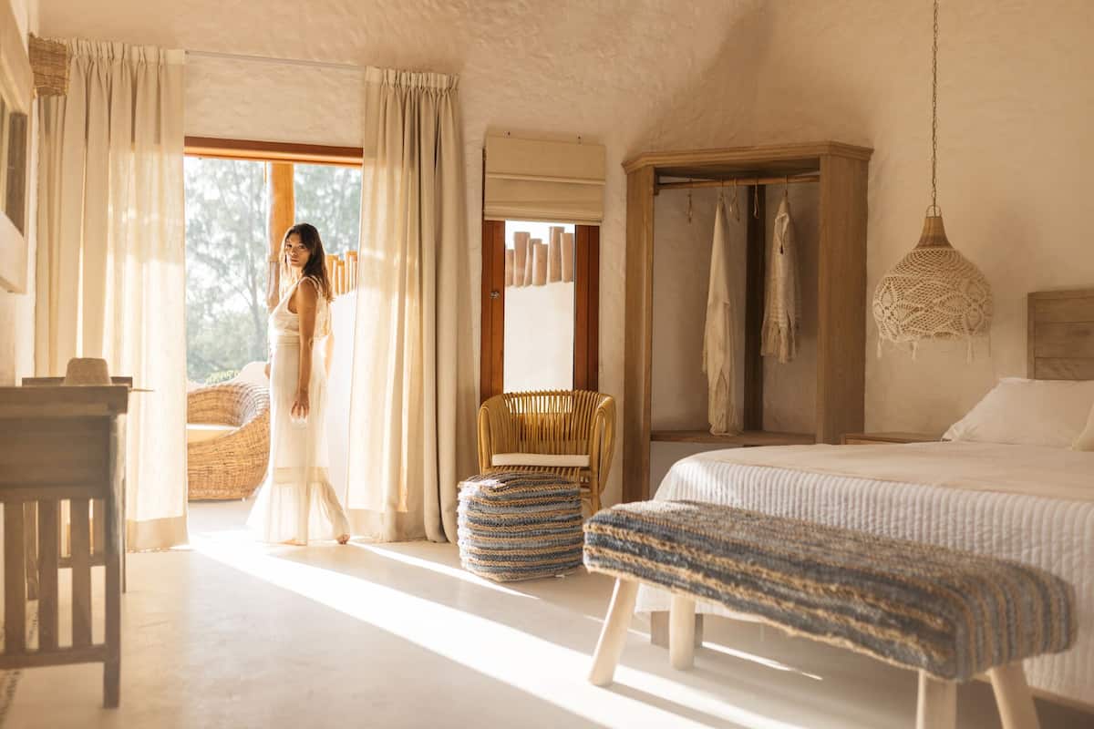 A bedroom with a bed, a bench, a chair, a hanging light fixture, and a window with curtains.