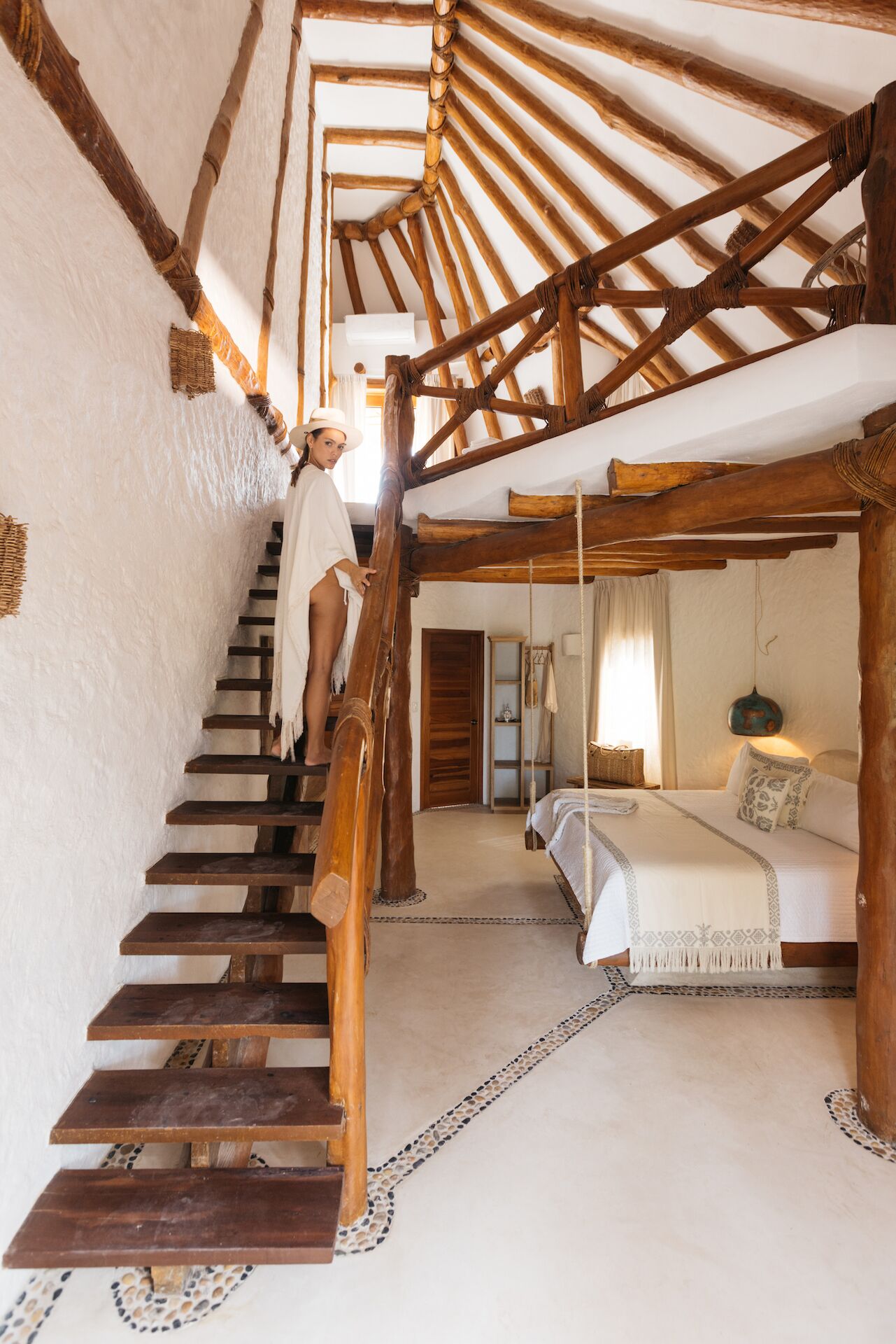A rustic wooden staircase leading to a bedroom with a bed, a hanging light fixture, and a small table.
