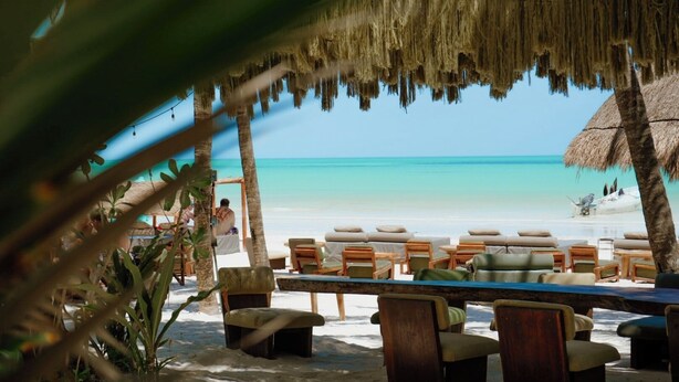 Un salón junto a la playa con techo de paja, mobiliario de madera y vista al océano.