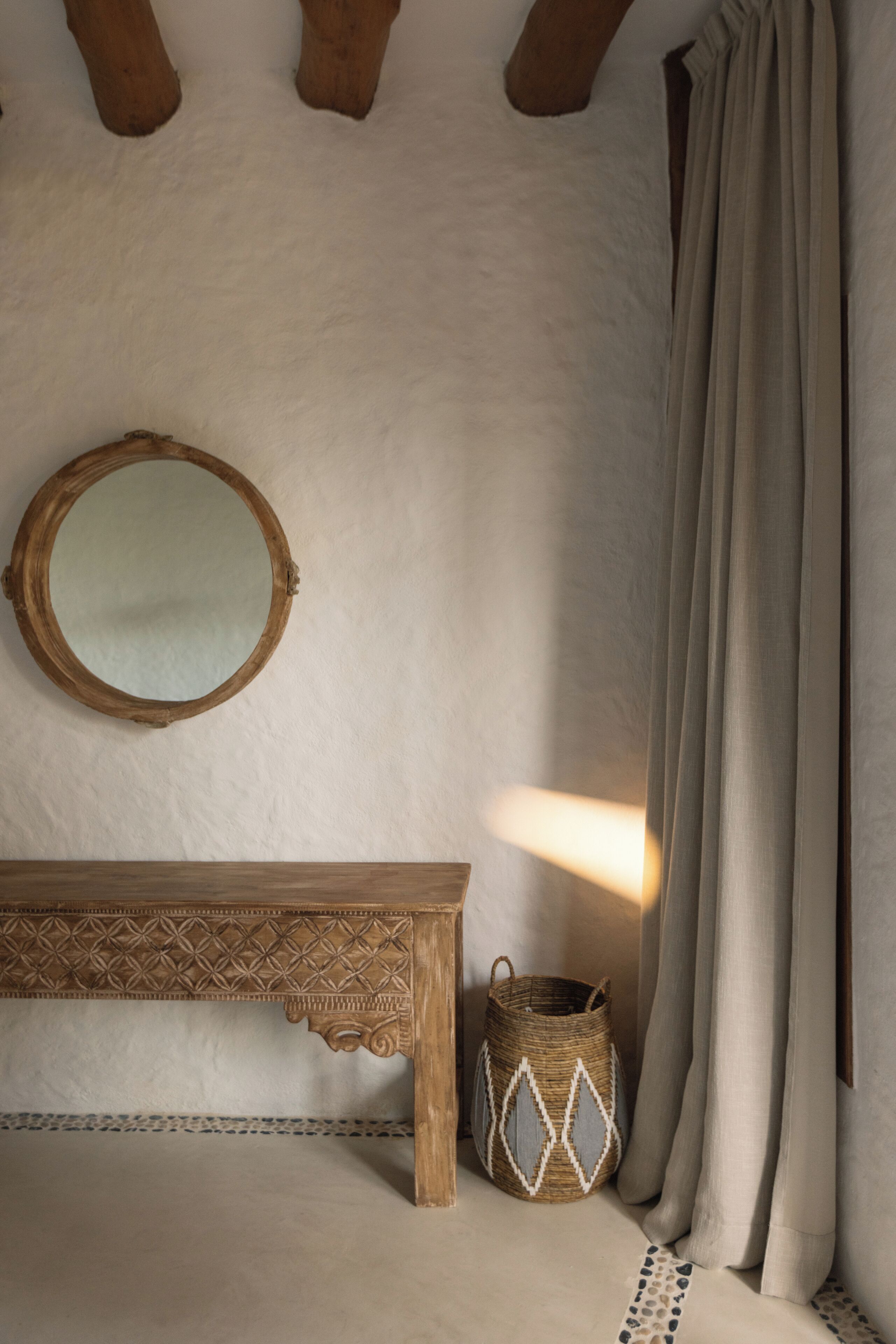 Una habitación con un banco de madera, una cesta tejida, un espejo redondo y una cortina.