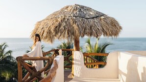 Un entorno de playa con un toldo de paja, barandillas de madera y una palmera al fondo.
