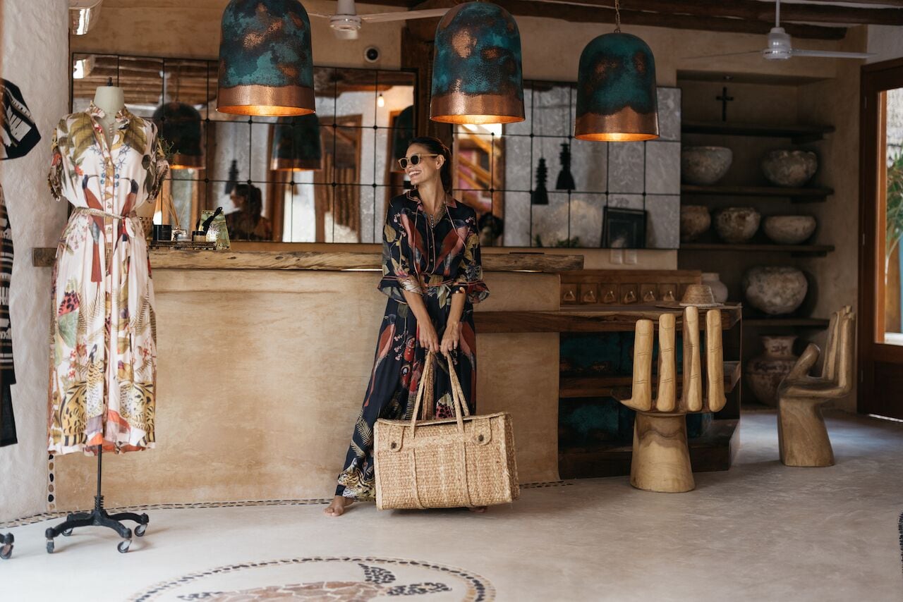 Interior de um hotel boutique com uma mesa de recepção de madeira, um manequim vestido com um vestido floral e uma mulher de vestido estampado segurando uma bolsa tecida.