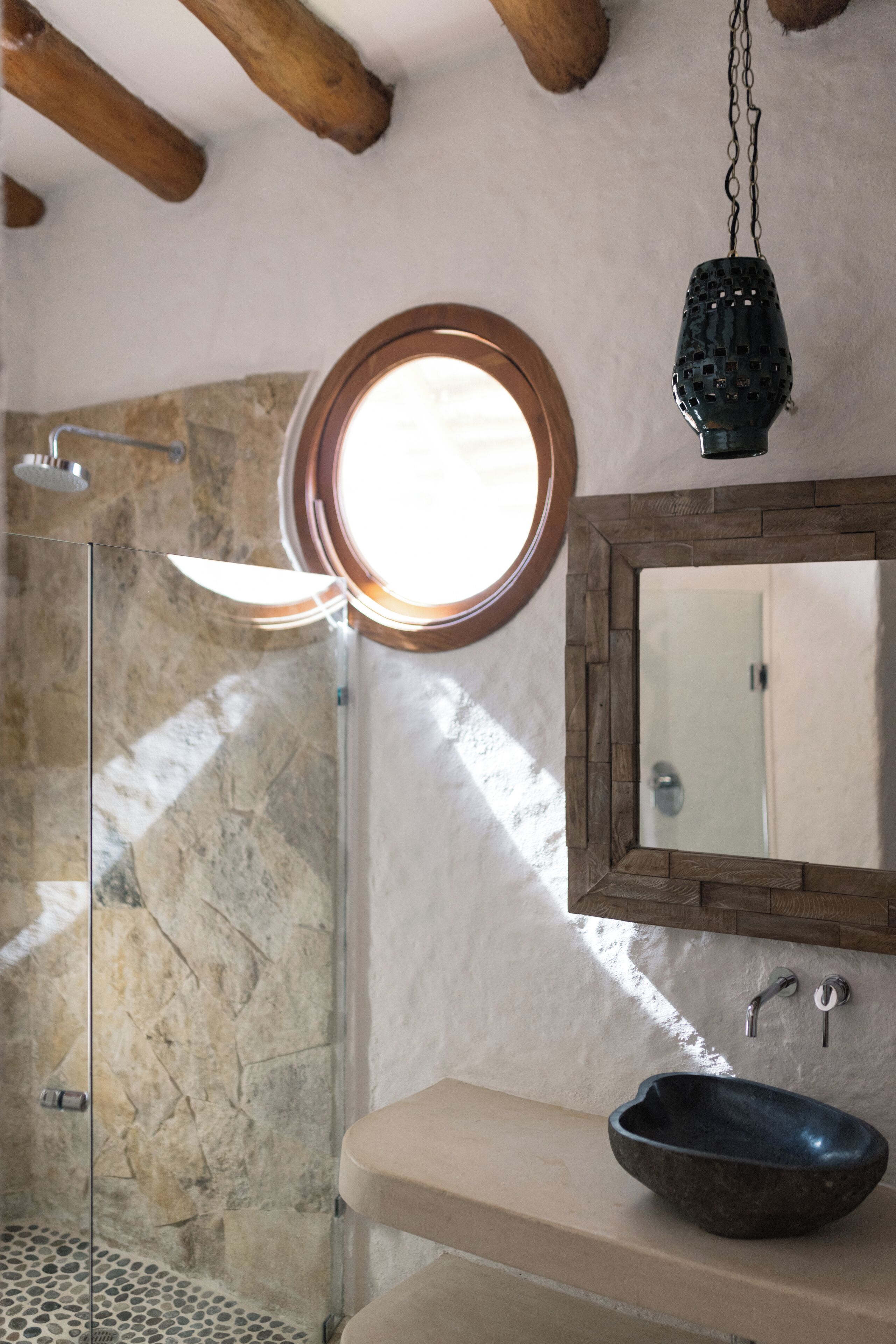 A bathroom with a stone shower, a round mirror, and a vessel sink.