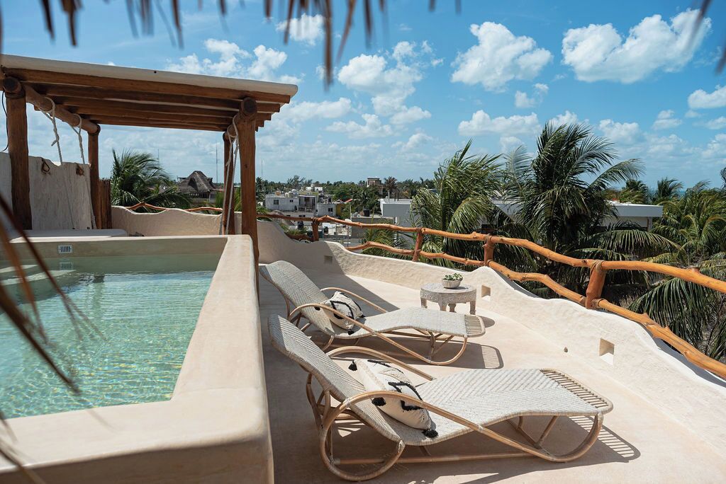 A poolside area with lounge chairs and a small table, surrounded by palm trees and a clear sky.