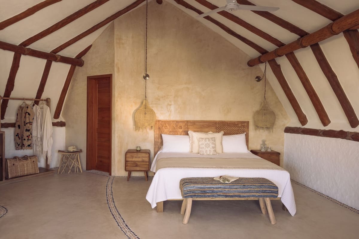 A bedroom with a wooden ceiling, a bed with white linens, two bedside tables, and a woven bench.