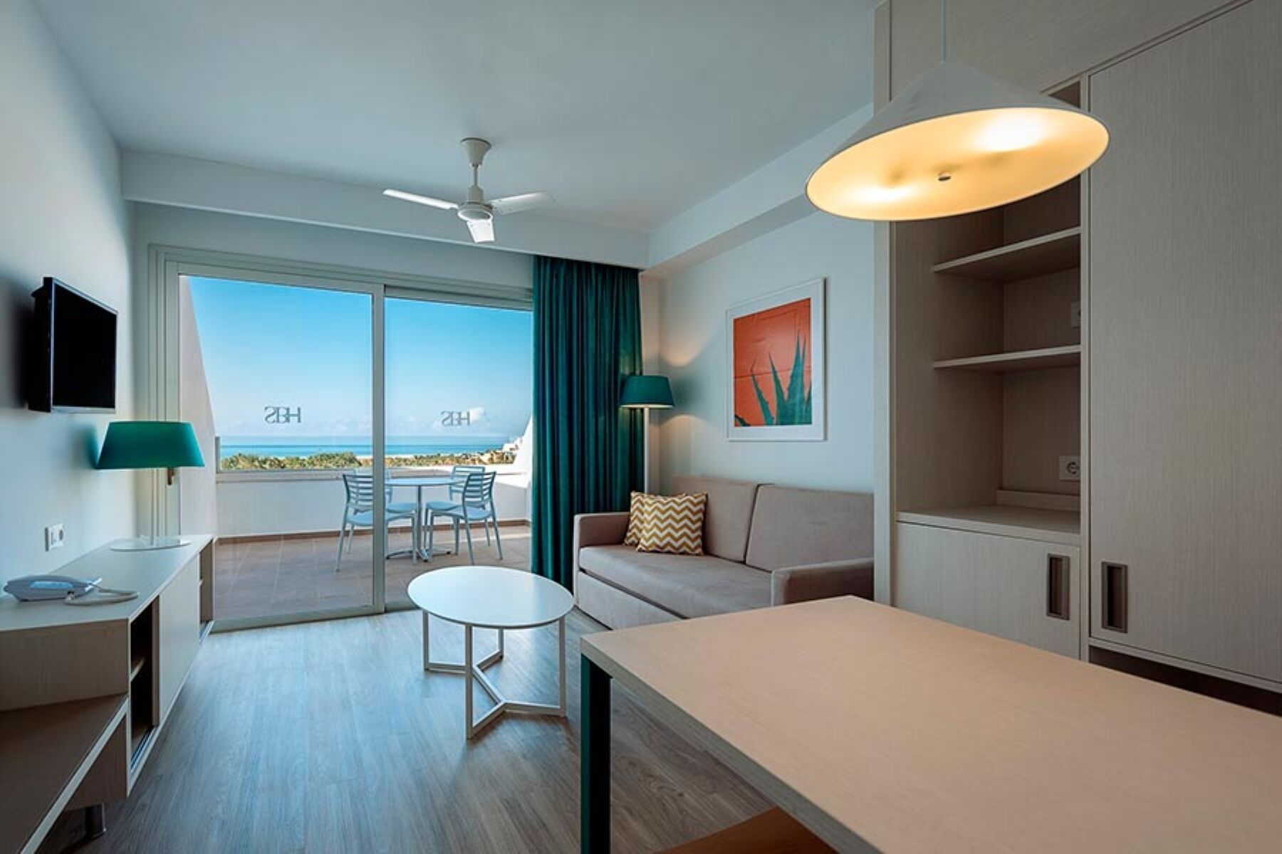 A modern hotel room with a balcony, a ceiling fan, a table, a sofa, and a TV.