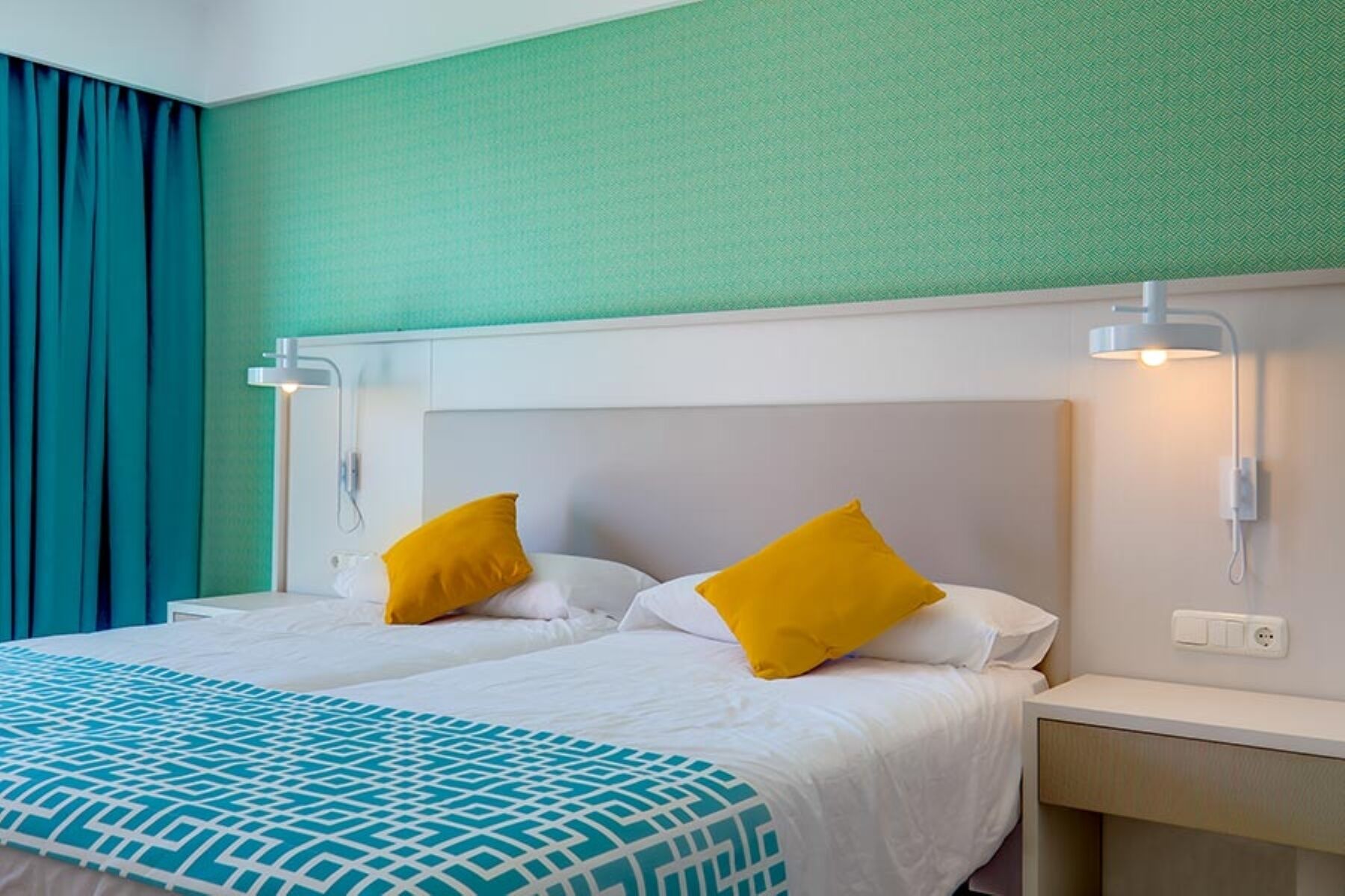 A hotel room with a bed, two bedside tables, a nightstand, and a window with curtains.